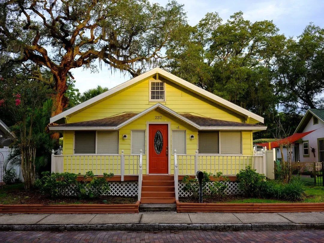A yellow craftsman bungalow with a red door and a covered front porch under an oak tree